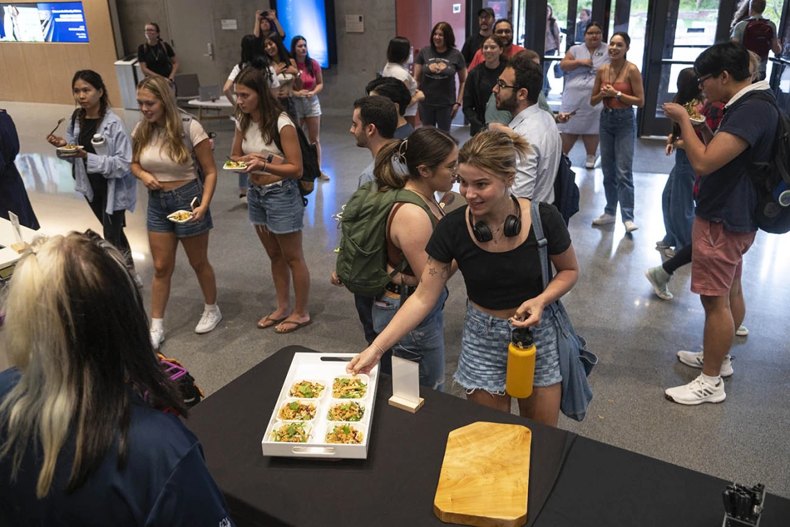 A young woman reaches for a small salad plate from a counter as dozens of other people mingle in the background. 