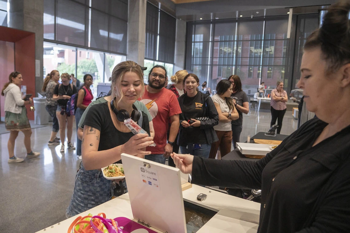 A young woman reaches for a small salad plate from a counter as dozens of other people mingle in the background. 