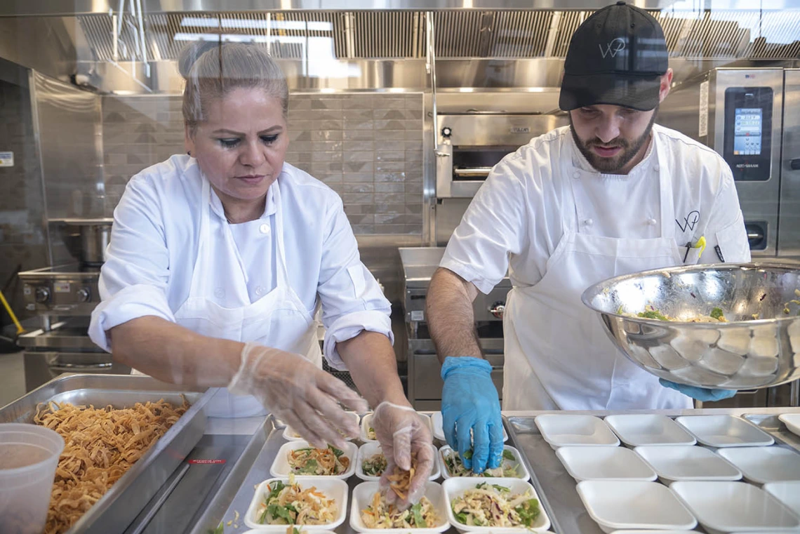 Two chefs in white coats prepare small salad plates in a kitchen. 