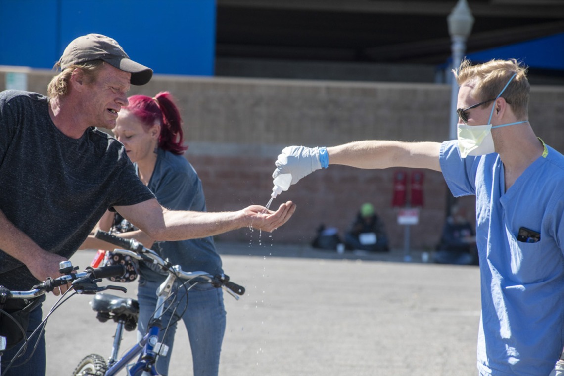 A student in the College of Medicine – Tucson helps a man sanitize his hands. Students are helping homeless people during the pandemic, because the medically-vulnerable population is expected to be hit hard by the COVID-19 virus.