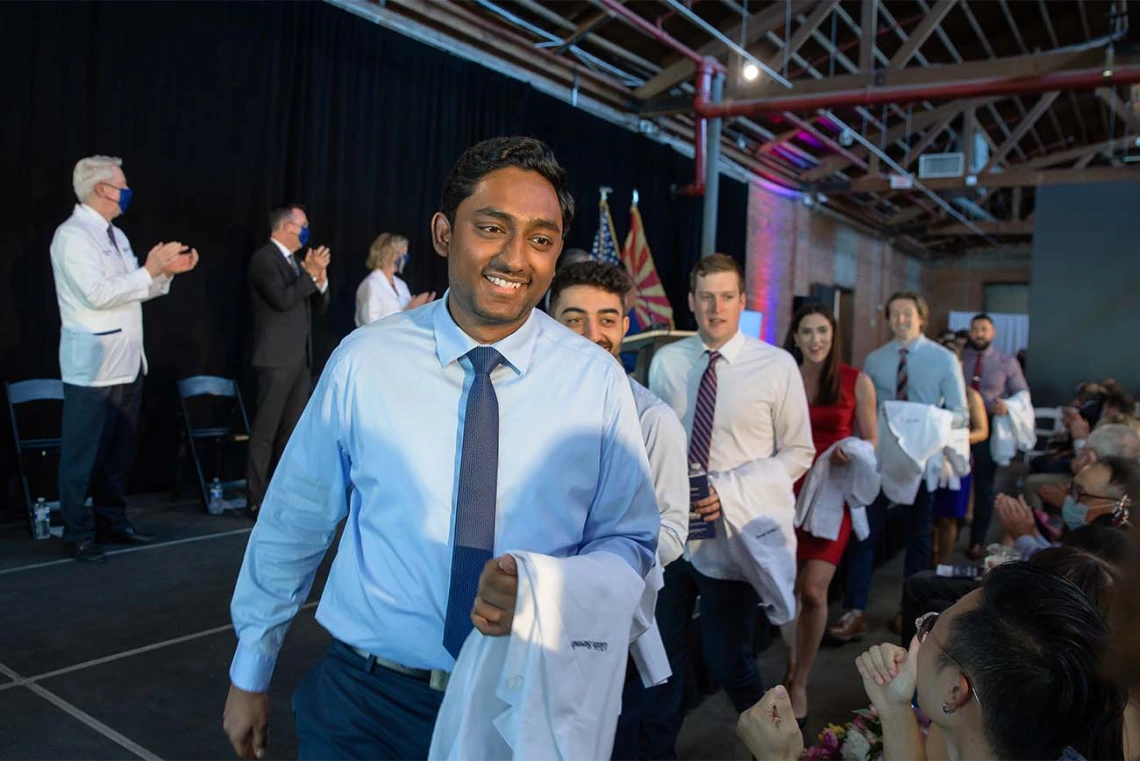 Lucky Surendra, Armin Talle, Gregory Sprout, Hannah Tolson, Tyler Vail and Ankedo Warda enter the white coat ceremony in front of seated friends and family.