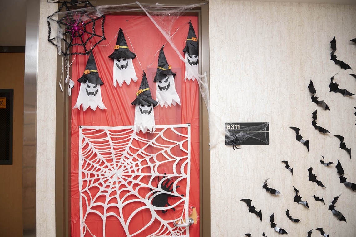 Door in a hallway covered in orange paper with several ghosts in witch's hats, a spiderweb and bats flying off on the wall to the side. 