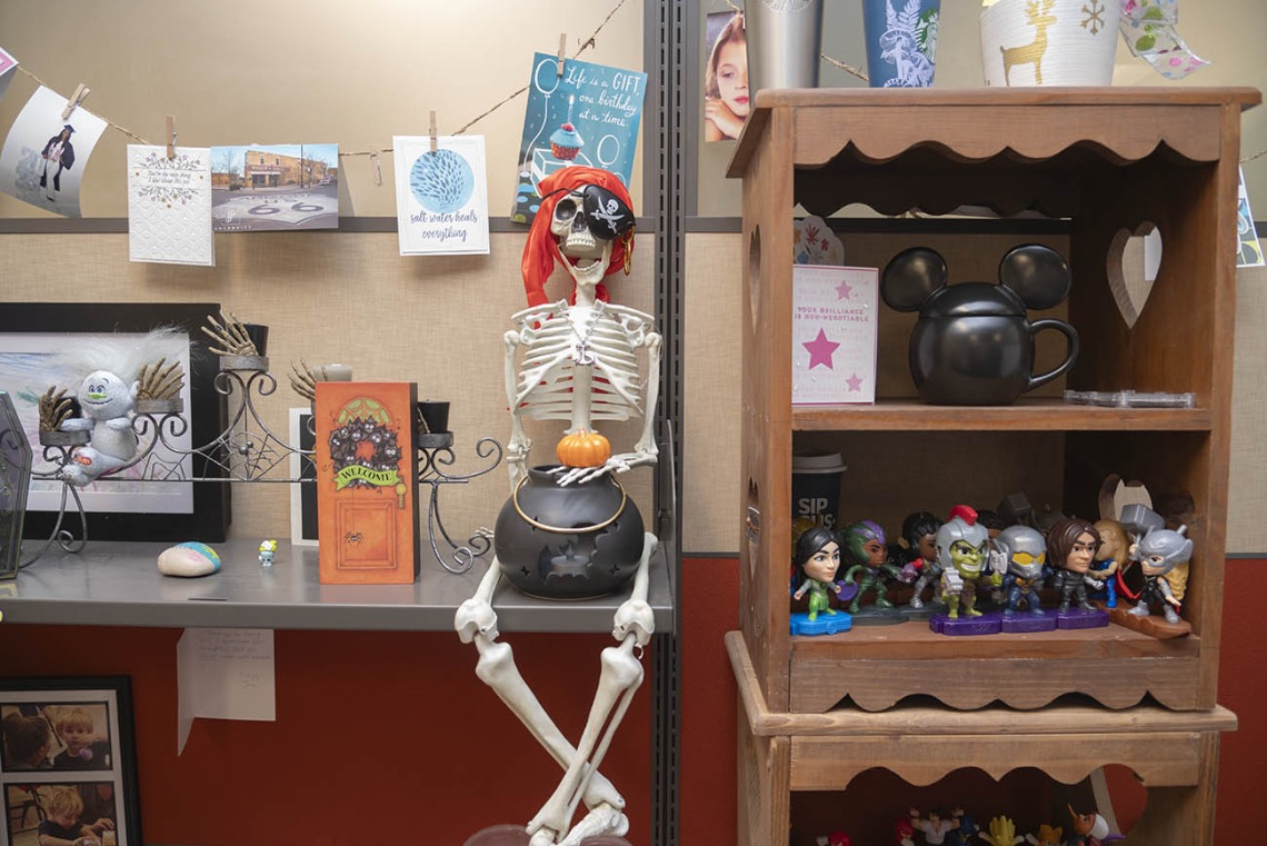 A skeleton with a pirate bandana on its head sits on a desk with a candy bowl between its legs. 