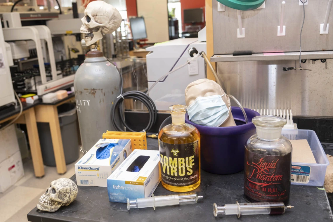 A laboratory table has larg jars labeled "Zombie Virus" along with a couple of skulls and a manikin head with a mask sitting in a bowl. 