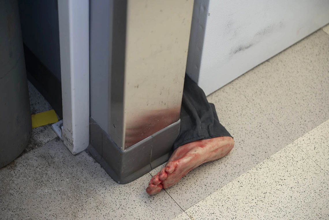 The lower part of a pantleg with a bloody foot sticks out from between a wall and piece of furniture. 
