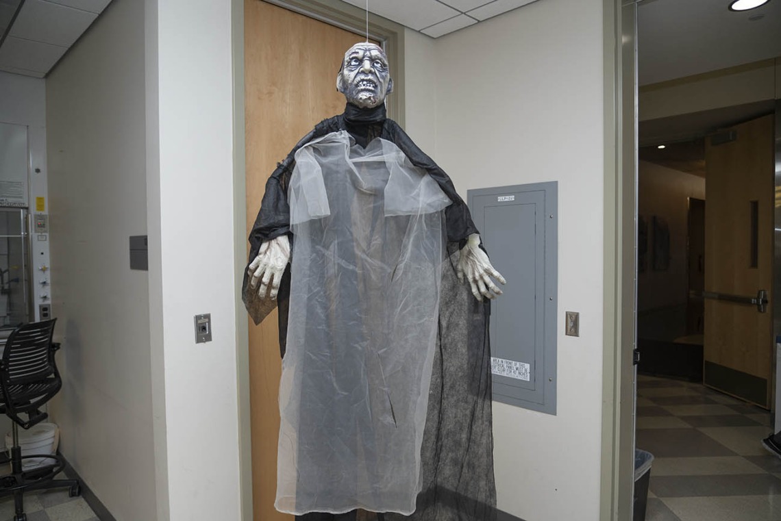A very tall zomby in a black robe stands in a hallway of a lab.
