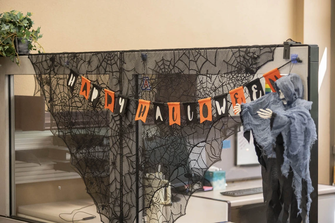 Black spider webs and an orange and black string of words say "Happy Halloween" along a cubicle wall. 