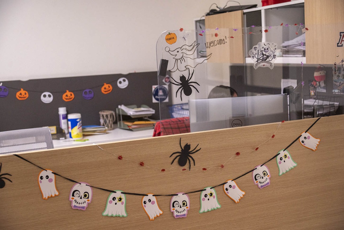 Little ghosts hang on a string across the front of a reception counter in an office. Spiders and small jack-o-lanters also decorate the area. 