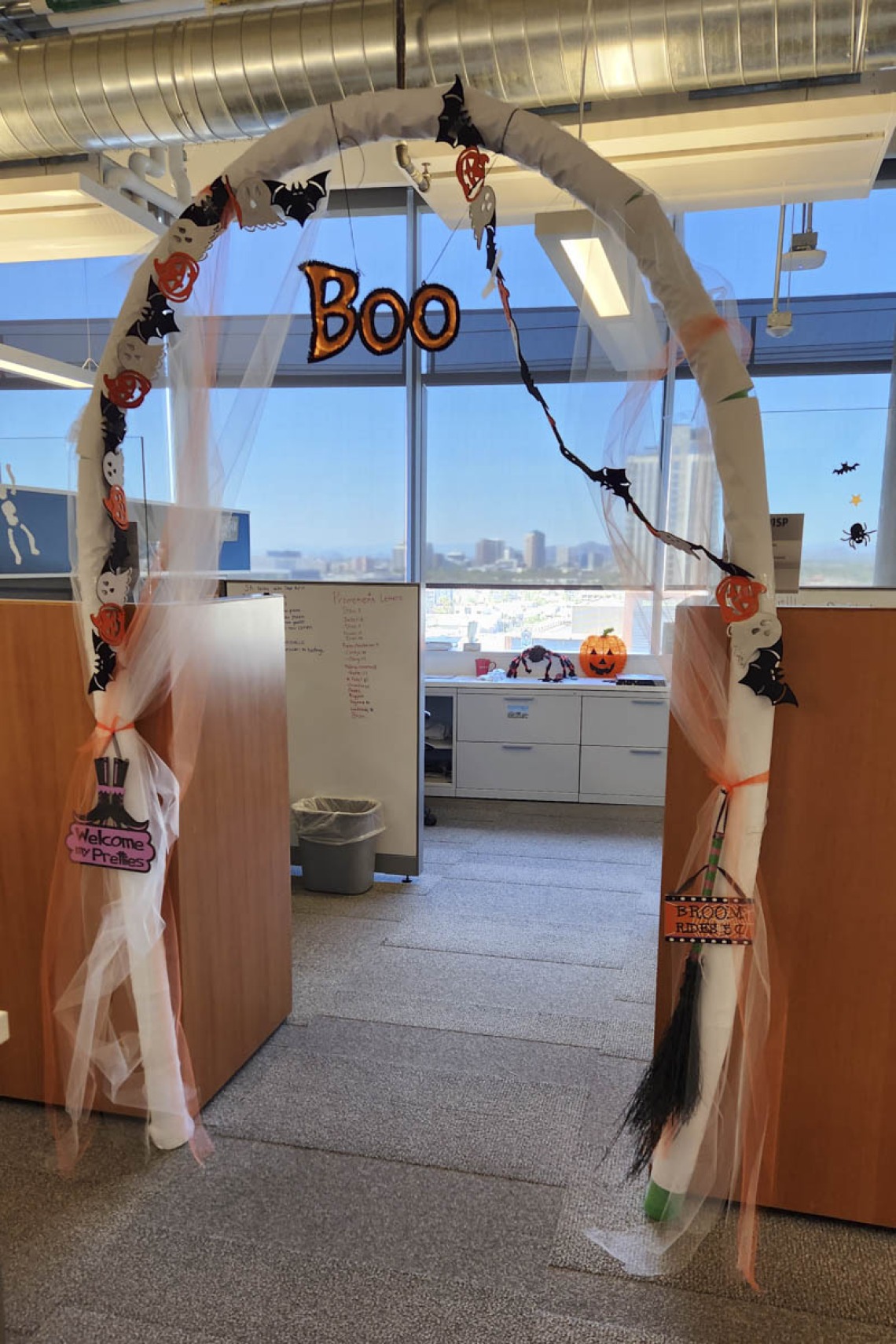 Halloween decorations create an archway between cubicles in an office. the word "Boo" hangs from the top.
