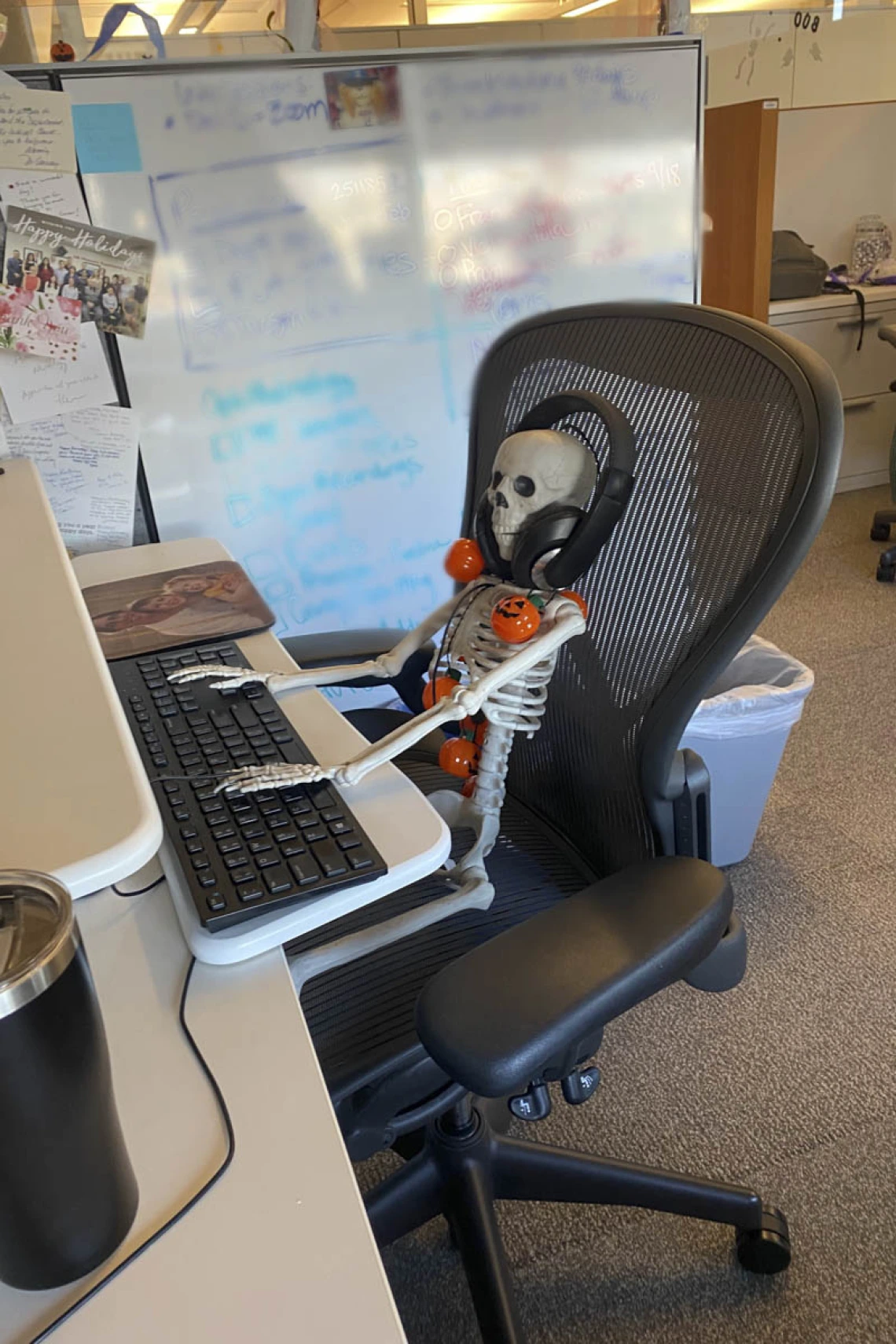 A skeleton sits in an office chair with hands on the computer keyboard and headphones on. 