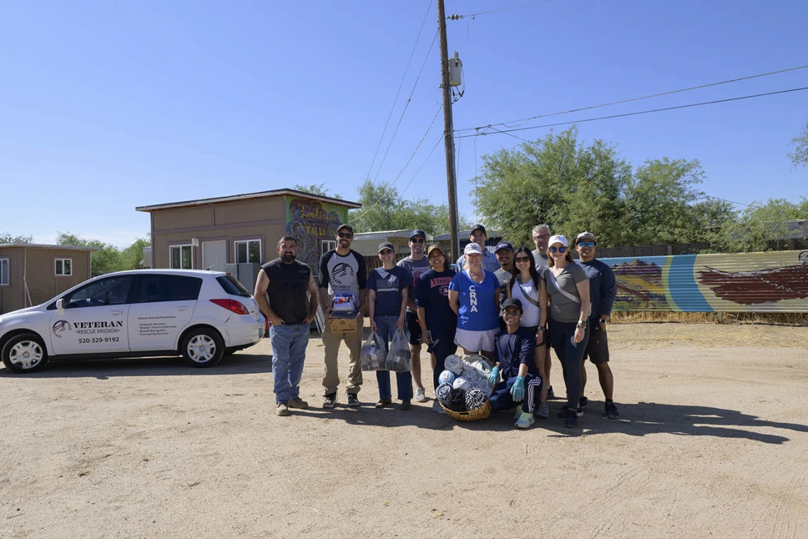 About a dozen people stand in a group in a dirt parking lot holding items they collected to donate to the Veteran Village. 