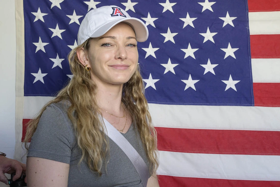 A young woman with long blonde hair and a white UArizona baseball hat smiles as she stands in front of an American flag on a wall. 