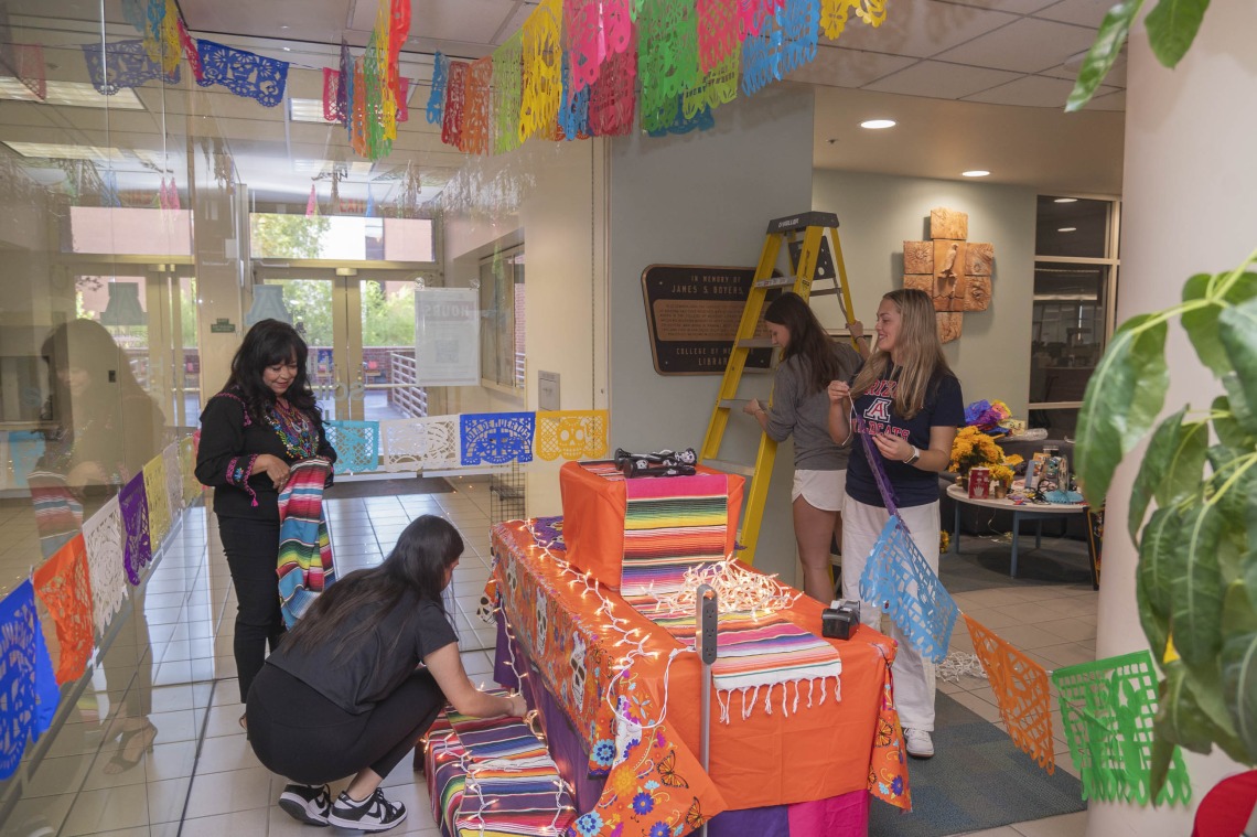 Four women work on setting up the Dia de los Muertos altar in the Health Sciences Library. They are hanging decorations using a ladder. 