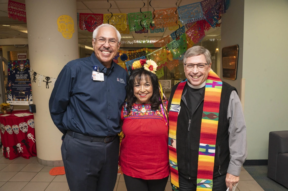 Two men stand on each side of a short woman, all smiling. They are in front of the Dia de los Muertos altar. 