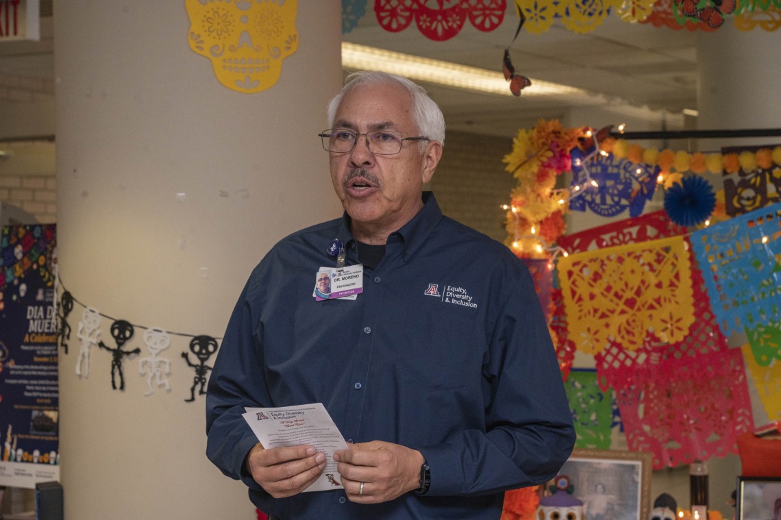Dr. Francisco Moreno, a Hispanic man with gray hair, holds a paper as he speaks in front of festivly decorated background. 