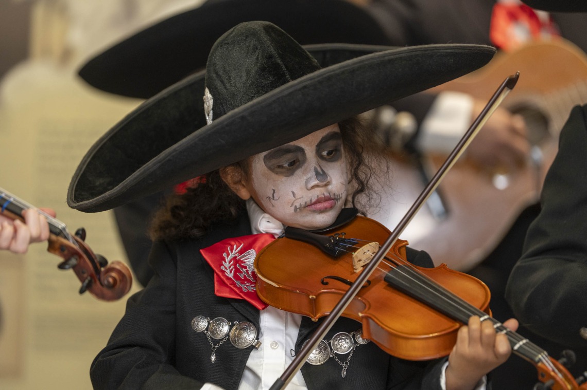 A young girl dressed in a black mariachi suit with a black sombrero hat plays a violin. Her face is painted in a traditional black and white skull design. 