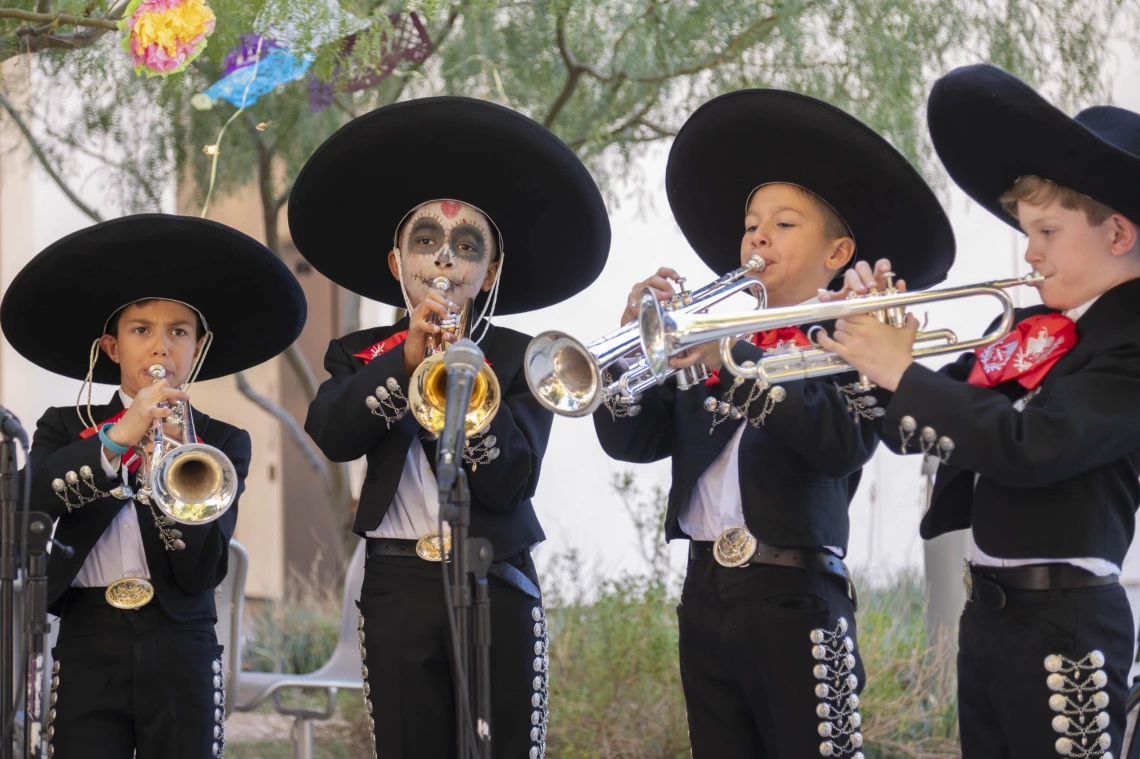 Four boys dressed in black mariachi suits with large brimmed black sombrero hats all playing trumpets.