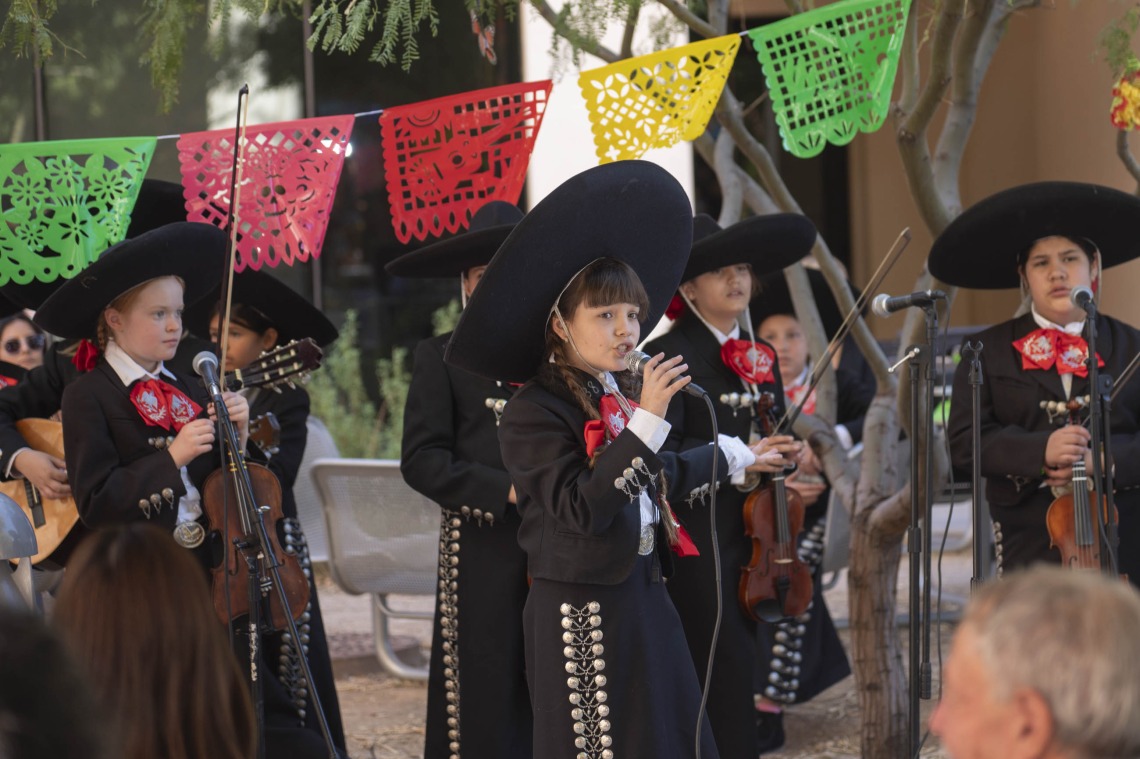 A young girl in a mariachi suit with large black sombrero sings into a microphone as severa kids in the same outfit stand around her with musical instruments. 