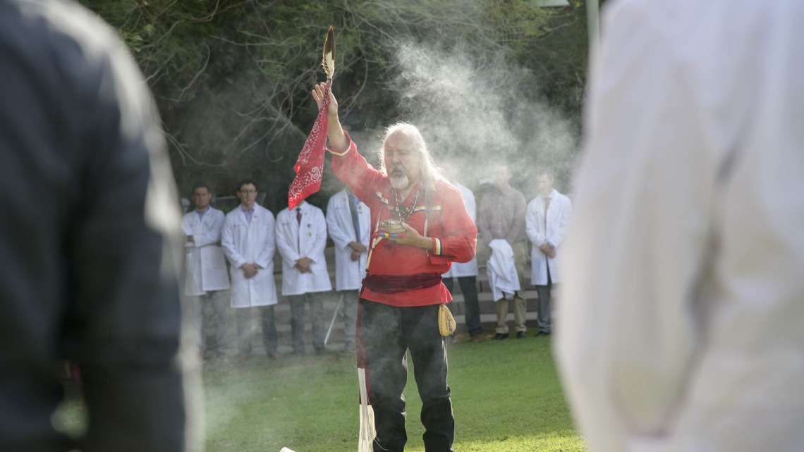 Native American man dressed in traditional tribal attire performs a ceremony surrounded by doctors dressed in white coats