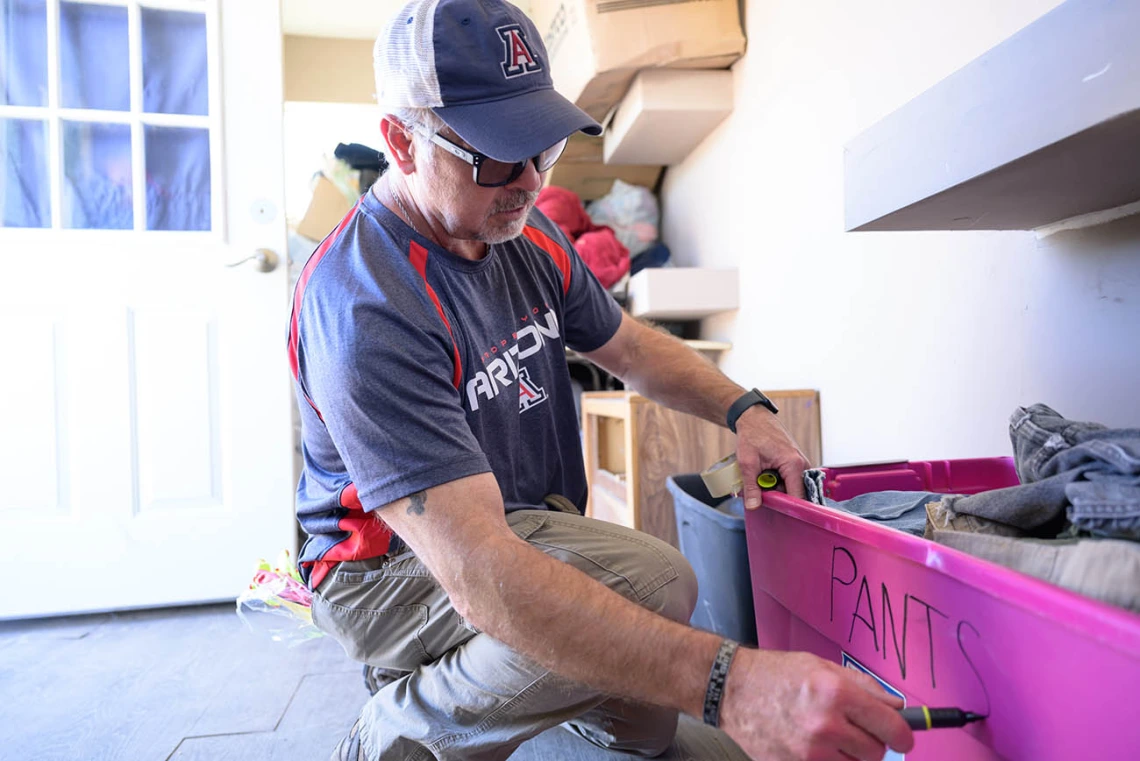 A white man squats down and writes "pants" onto the side of a pink tub full of pants. 