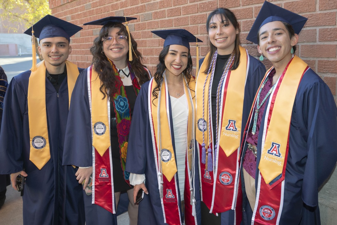 Five smiling nursing graduates in caps and gowns stand together outside. All have sashes on with the University of Arizona "A". 