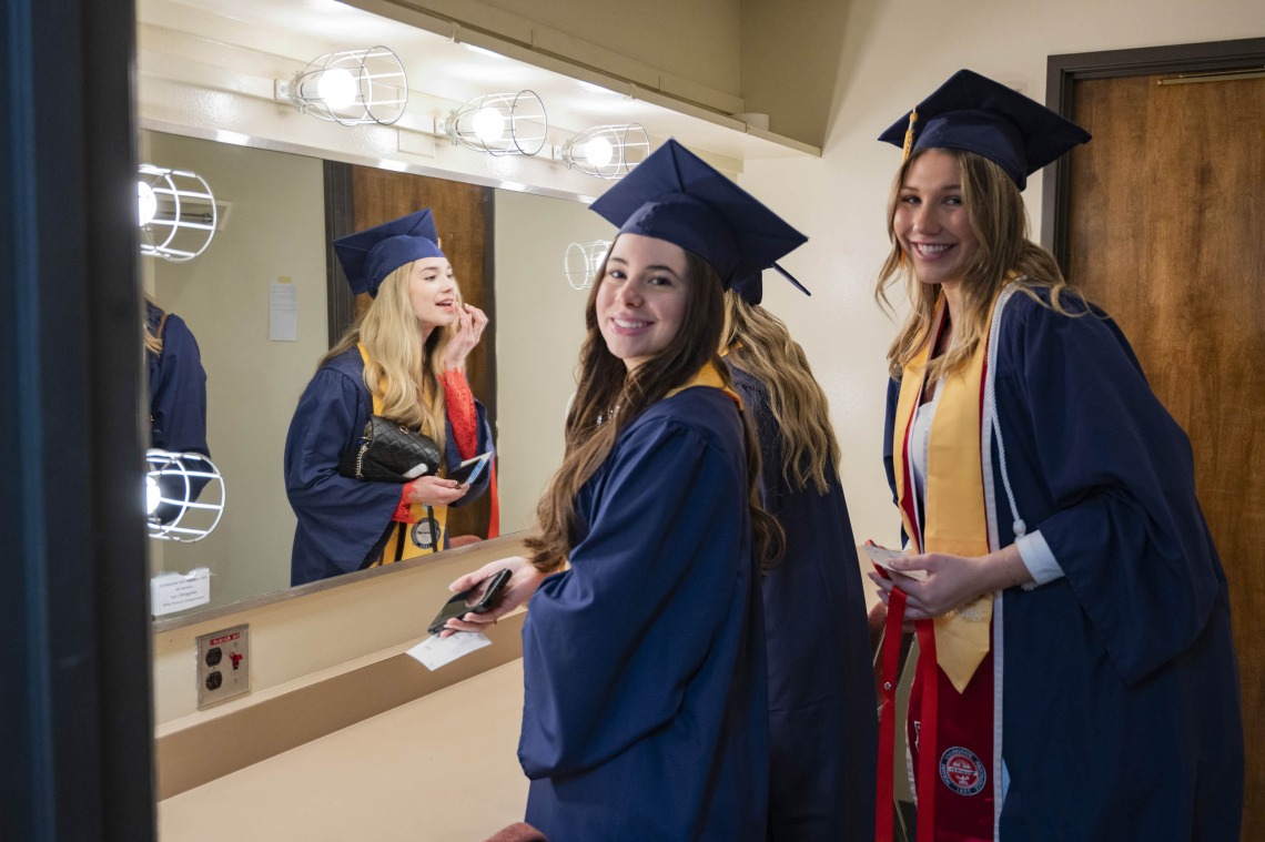 Three young women in graduation caps and gowns stand in a dressing room with a mirror. Two are smiling and one is putting lip gloss on in the mirror.