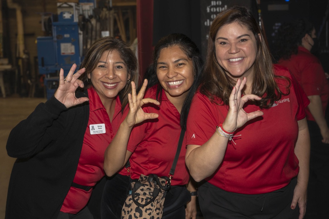 Three smiling women in red polo shirts all make the Arizona Wildcat sign with their fingers. 