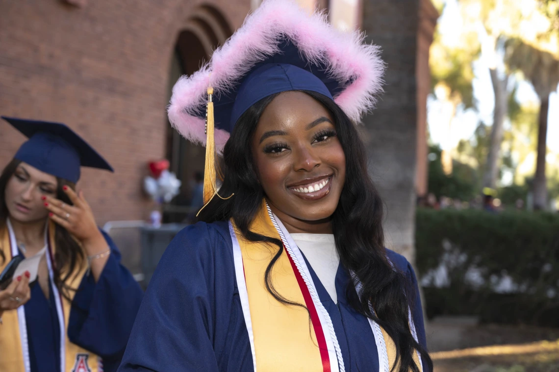 A young woman wearing a graduation cap decorated with pink feathers and a graduation gown smiles as she stands outside. 