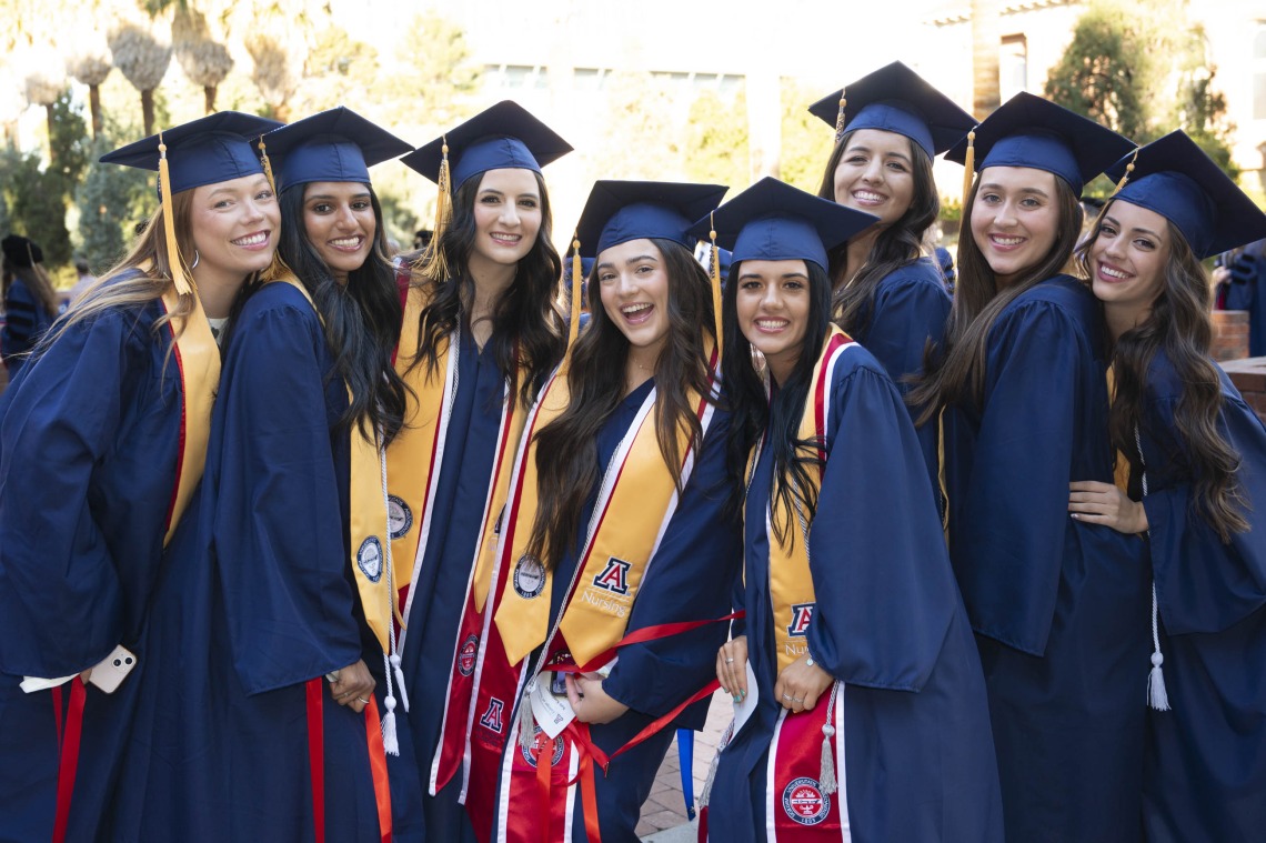 A groupl of eight young women stand together smiling, all dressed in graduation caps and gowns. 