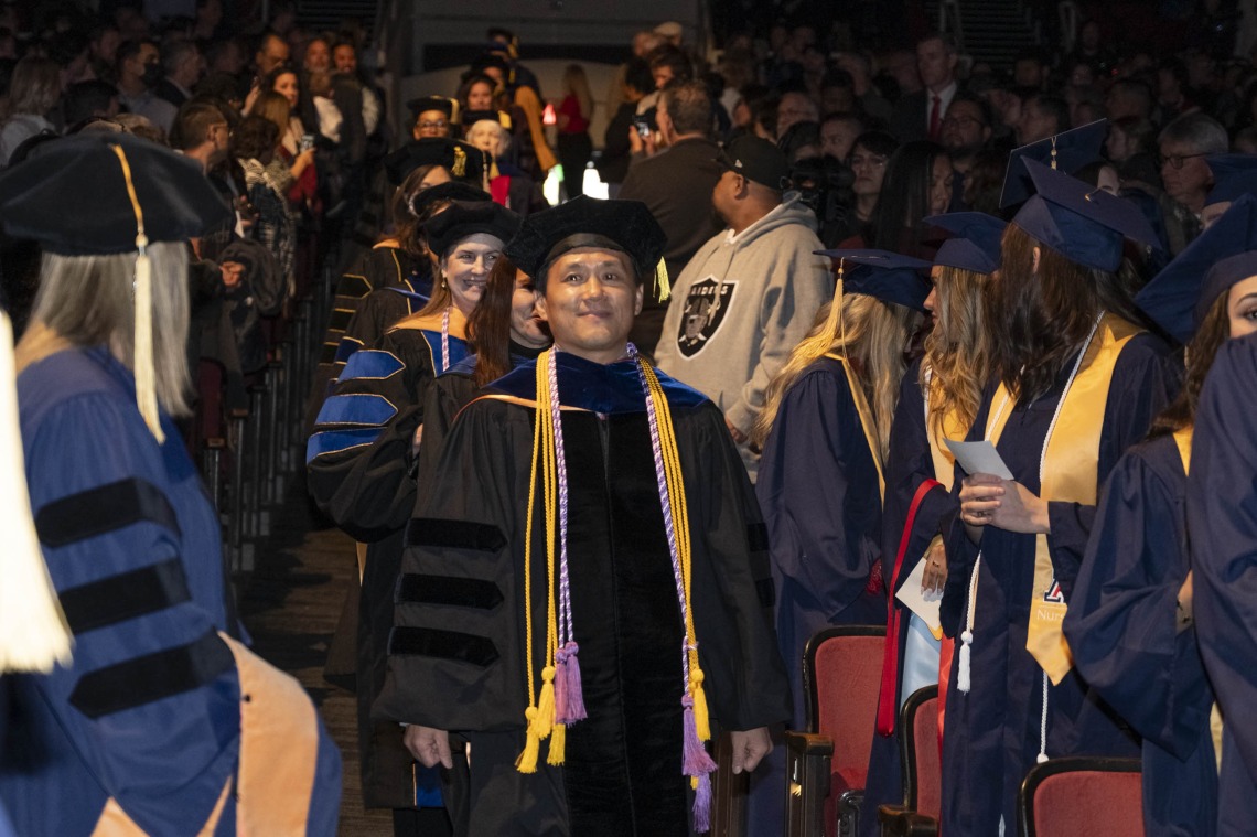 A line of nursing faculty walk down an aisle with students on both sides of them. All are wearing grauduation caps and gowns. 