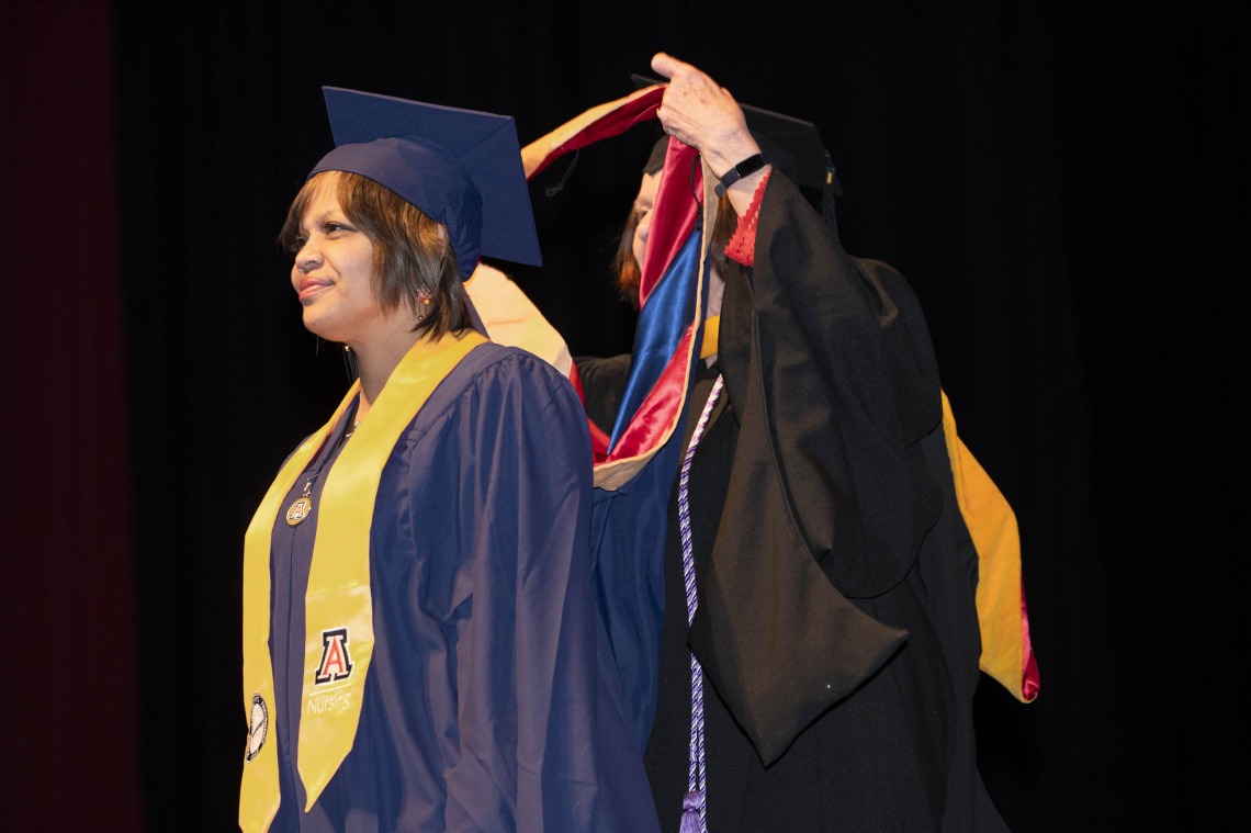 A female nursing student in graduation cap and gown stands looking forward as a professor places her graduation sash over her head. 