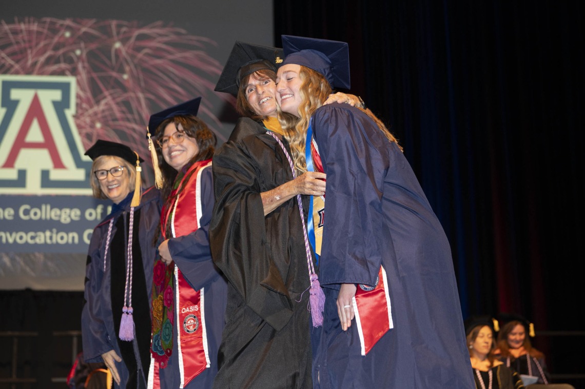 Two nursing students and two professors, all in graduation regalia, hug and smile on a stage. 