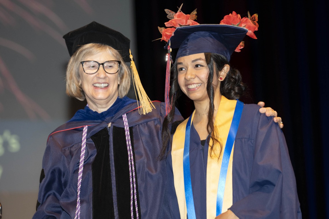A smiling professor and student stand together in their graduation regalia. 