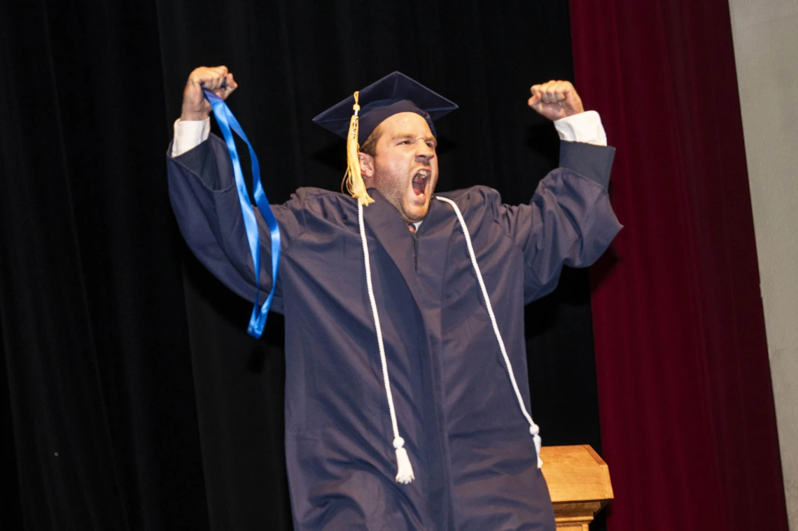 A young man wearing a graduation cap and gown raises both fists in the air and cheers. 