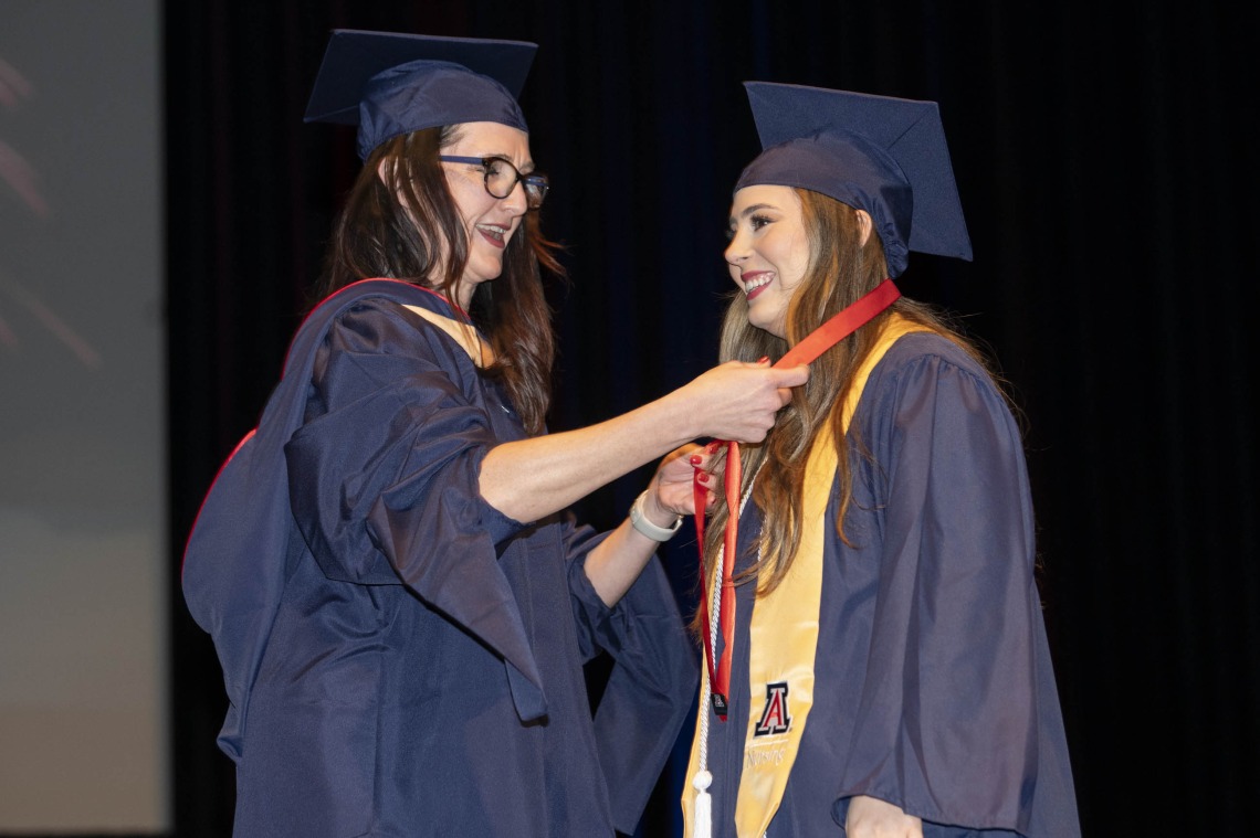 A nursing professor puts a lanyard on a nursing student. Both are wearing graduation caps and gowns. 