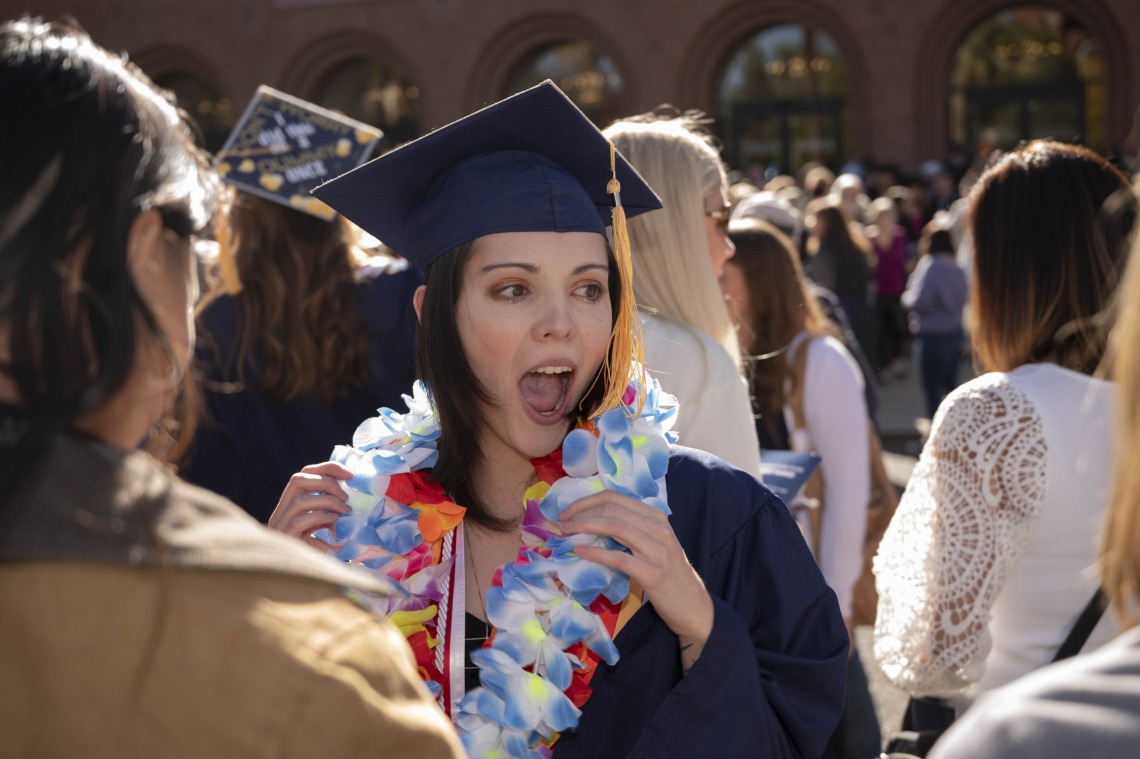 A young woman in a graduation cap and gown has her mouth open in a cheer in a crowd of people outside. 