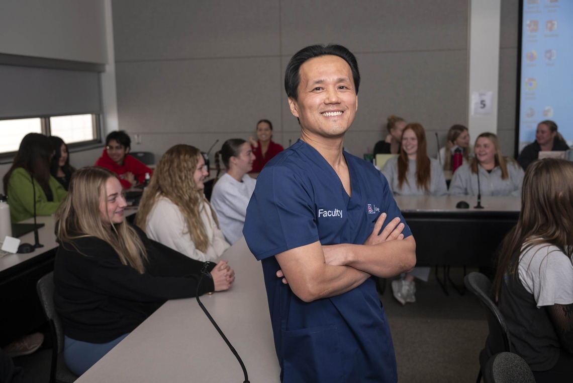Portrait of Dean Hyochol Brian Ahn wearing navy blue nursing scrubs and standing in front of a class of nursing students.