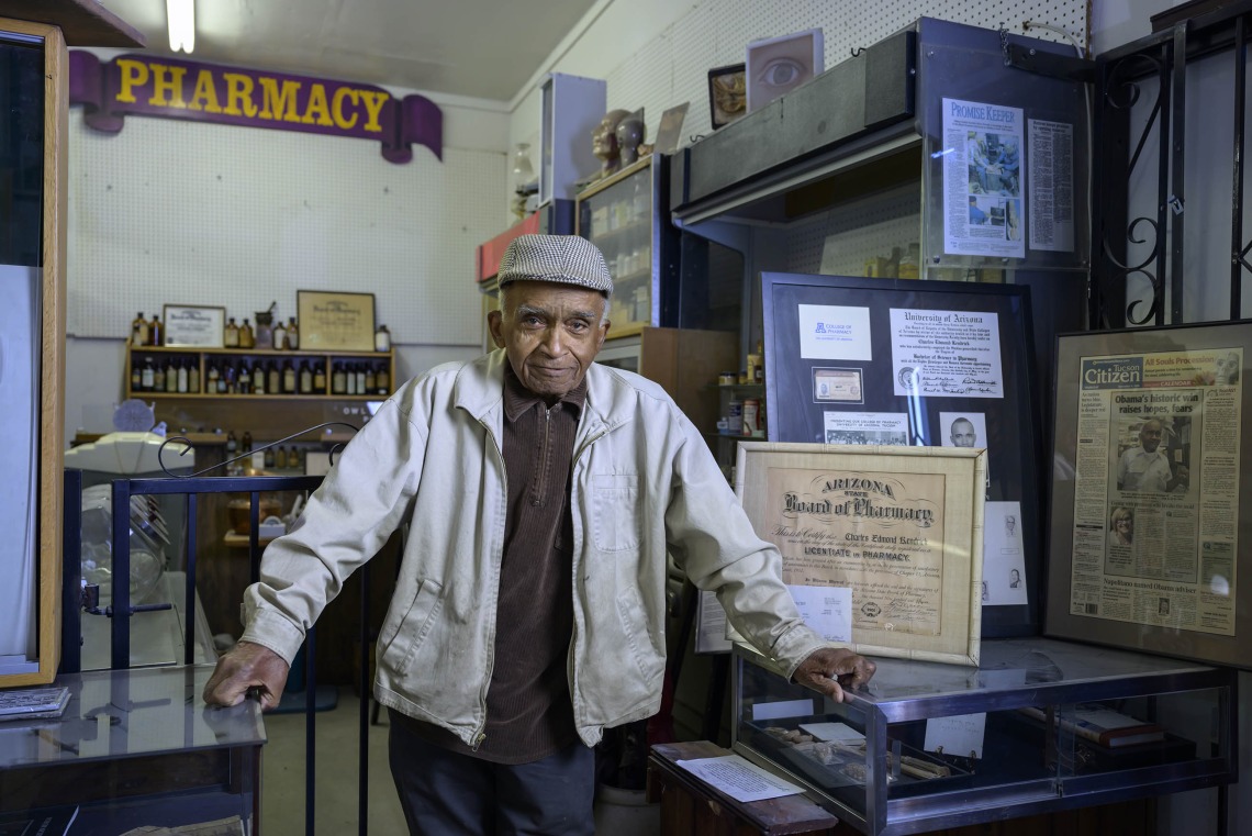 Charles Kendrick stands beneath a sign reading pharmacy and is surrounded with other pharmacy memorabilia.