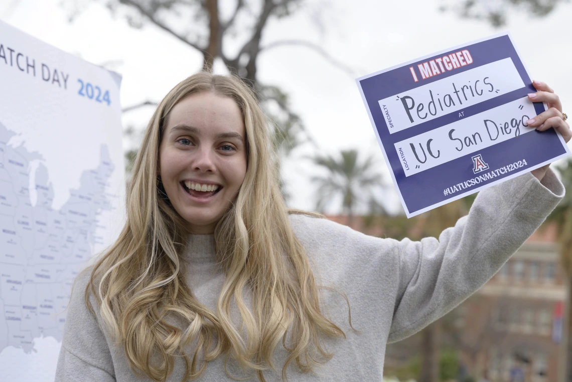 A smiling female, fourth-year medical student from the University of Arizona College of Medicine – Tucson holds a sign that says “I Matched” as she stands next to a map of the United States.