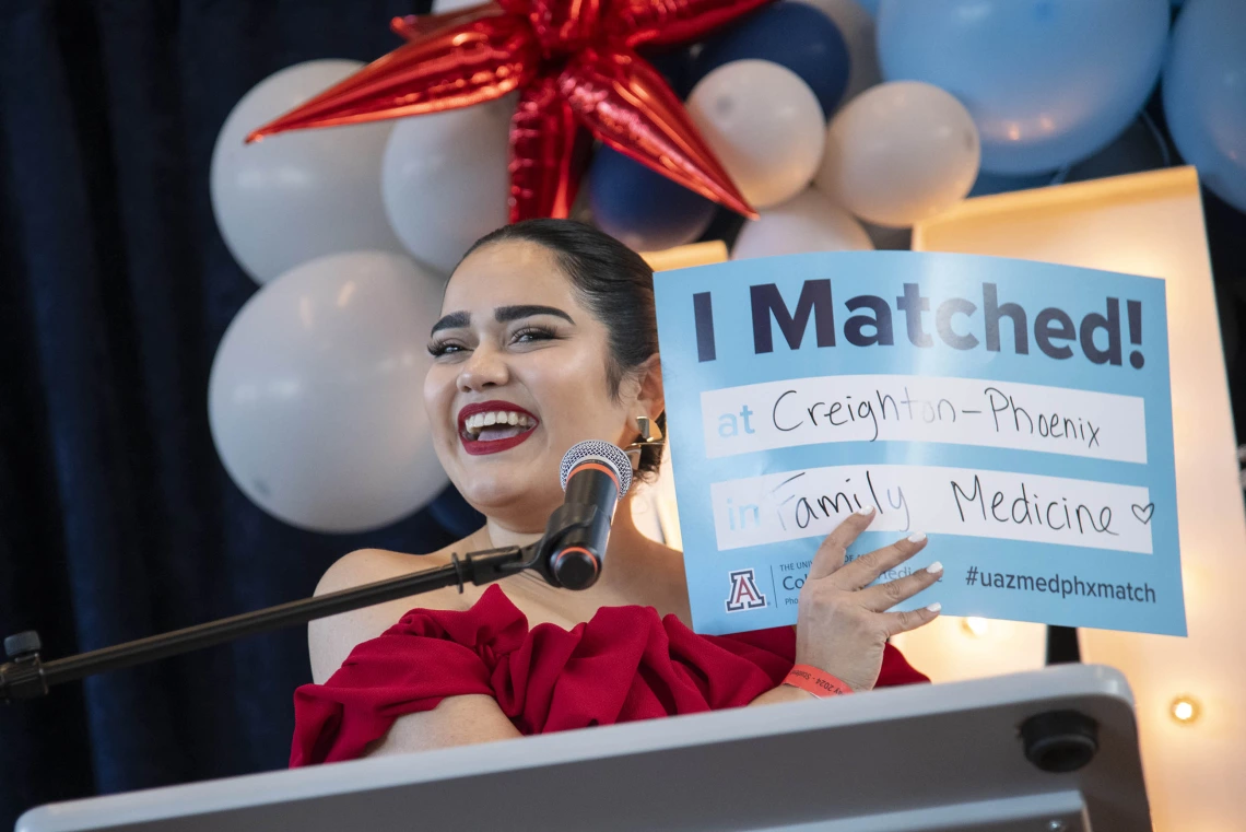 A smiling female, fourth-year medical student from the University of Arizona College of Medicine – Phoenix stands at a podium holding up a sign that says “I Matched” as she announces where she will be conducting her residency.