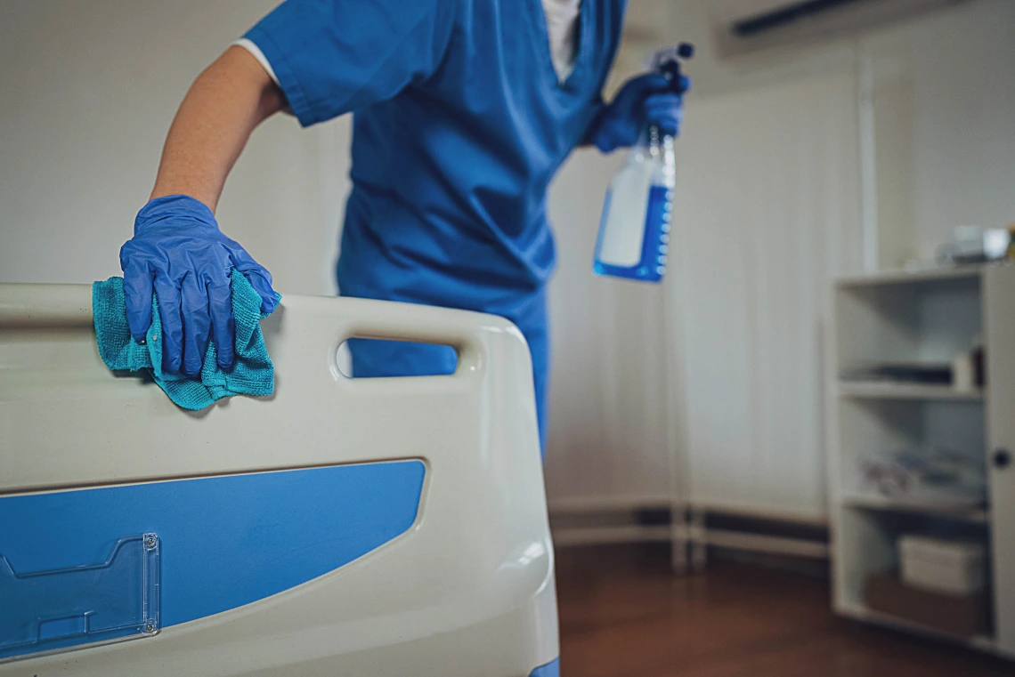 A nurse in blue scrubs wipes down a hospital bed with one hand while holding a spray bottle in the other.