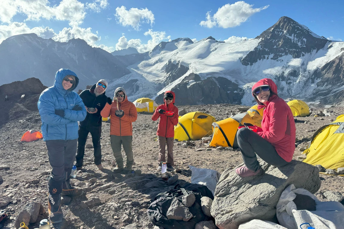 Snow-covered mountains are in the background as five men and women, all dressed in parkas, hold bowls of food.