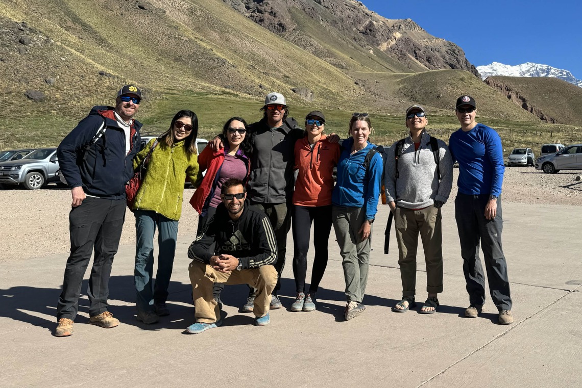 Nine people stand in a parking lot smiling with green mountains in the background. 