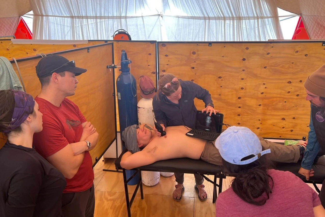 A man lies on his back on a table while Nicola Baker holds a portable ultrasound machine on his stomach in one hand and an ultrasound wand in another hand against his chest. 