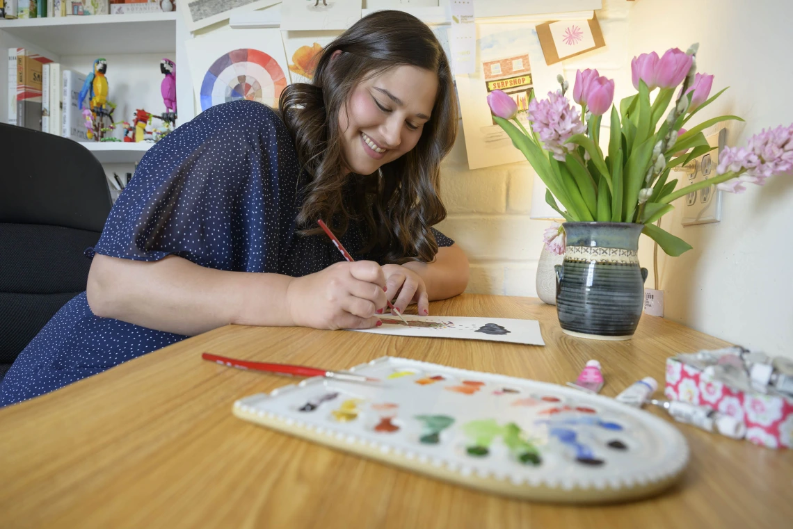 Anna Alkozei smiles as she holds a watercolor brush and paints a picture. A paint palette is in front of her and a vase of tulips in the background.