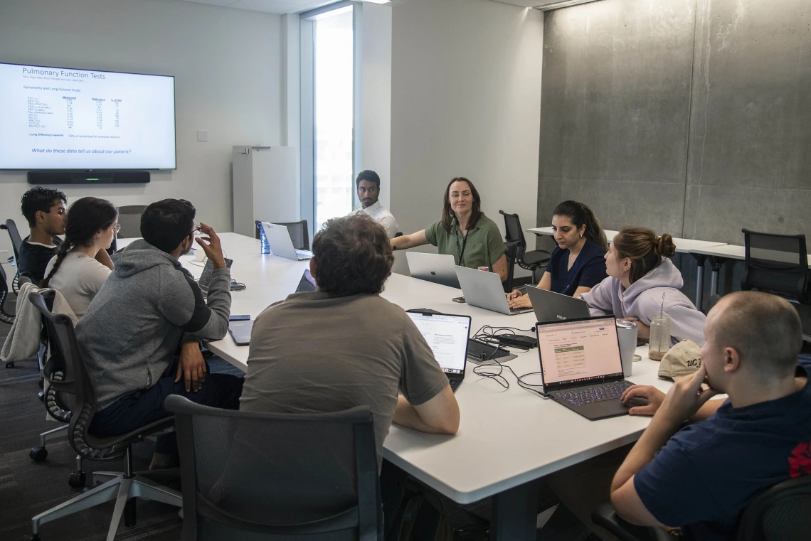 Halszka Glowacka sits at a conference table surrounded by students with a screen with writing on it in the background.