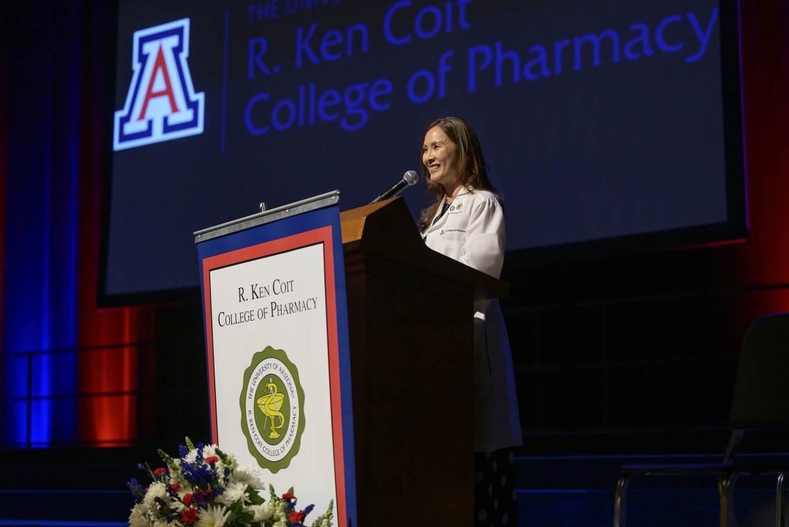 Professor Jeannie K. Lee speaks at a podium on a stage with the University of Arizona logo and “R. Ken Coit College of Pharmacy” projected on a screen behind her. 