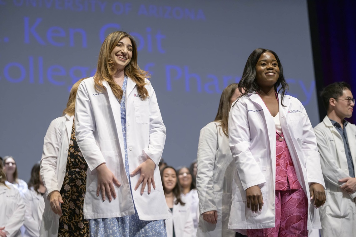Two University of Arizona College of Pharmacy students smile as they stand in front of other students, all wearing their white coats. 