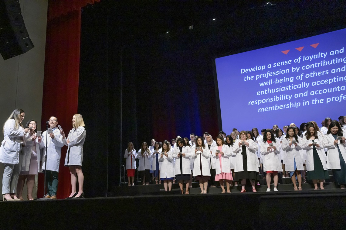 Four University of Arizona R. Ken Coit College of Pharmacy students stand around a microphone on the left while dozens stand in the background, all reading a professionalism pledge from cards they are holding.