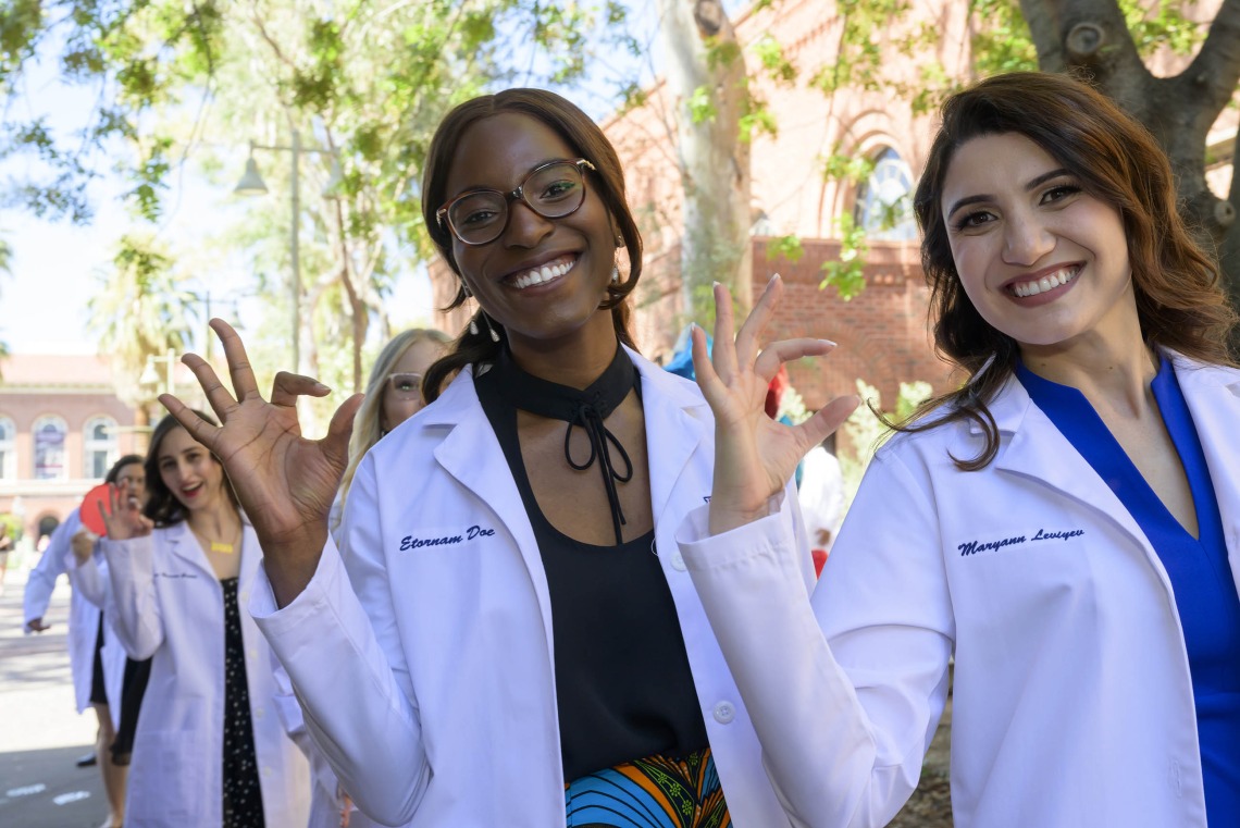 Two UArizona R. Ken Coit College of Pharmacy students smile and make the Wildcat sign with their fingers as they walk outside after receiving their white coats at a ceremony.